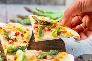 Stück Flammkuchen mit Schinken und grünem Spargel in der Hand