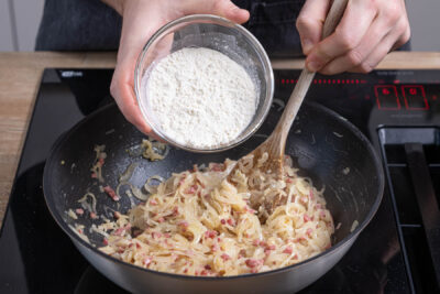 Schinkenwürfel, Mehl und Milch-Ei-Mischung zu den Zwiebeln geben