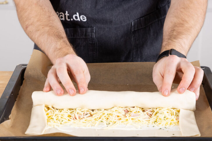 Den Blätterteigstrudel mit Schinken und Käse von unter her aufrollen.