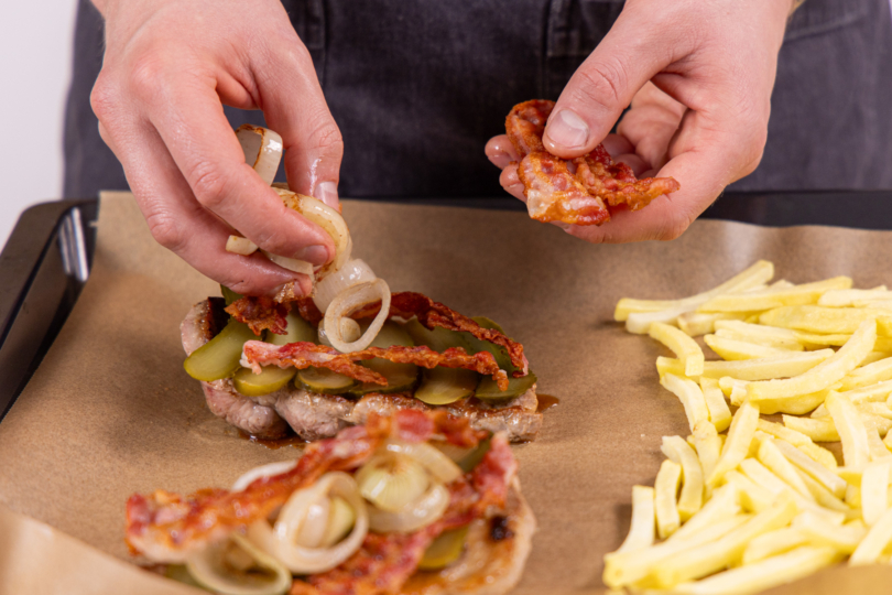 Bacon, Gurken und Zwiebeln auf den Steaks verteilen