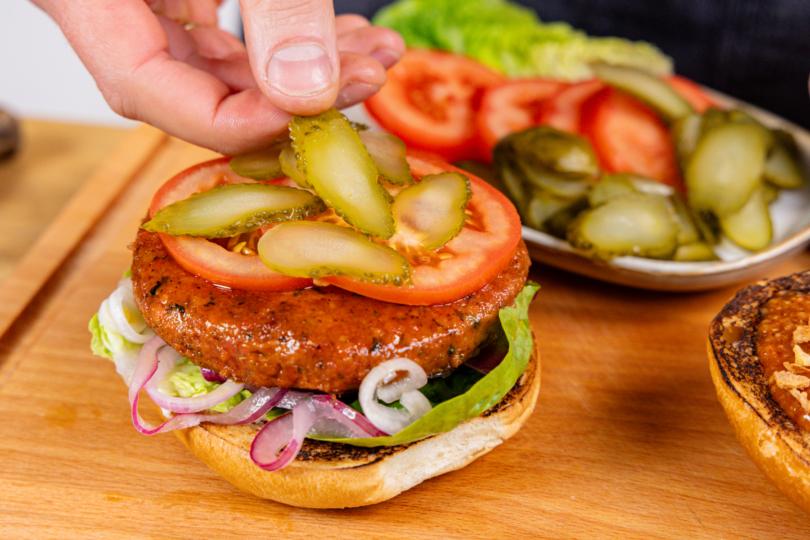 Tomaten und Gewürzgurken auf "Kein Burger"-Patty von Die Vegane Fleischerei platzieren